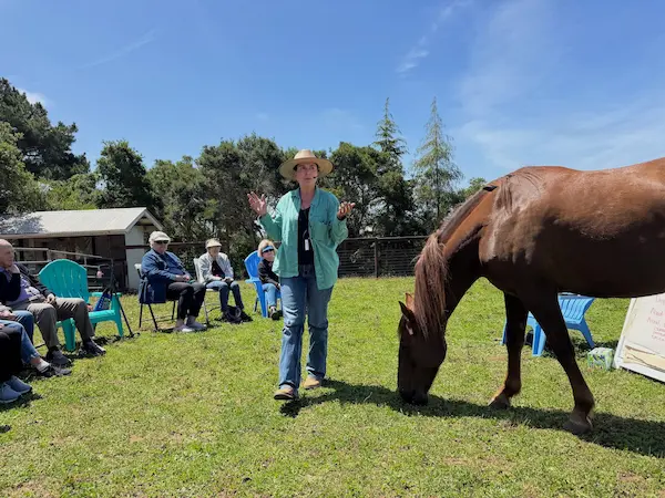 Woman presenting to a seated group outdoors with a horse nearby during a PMR educational workshop